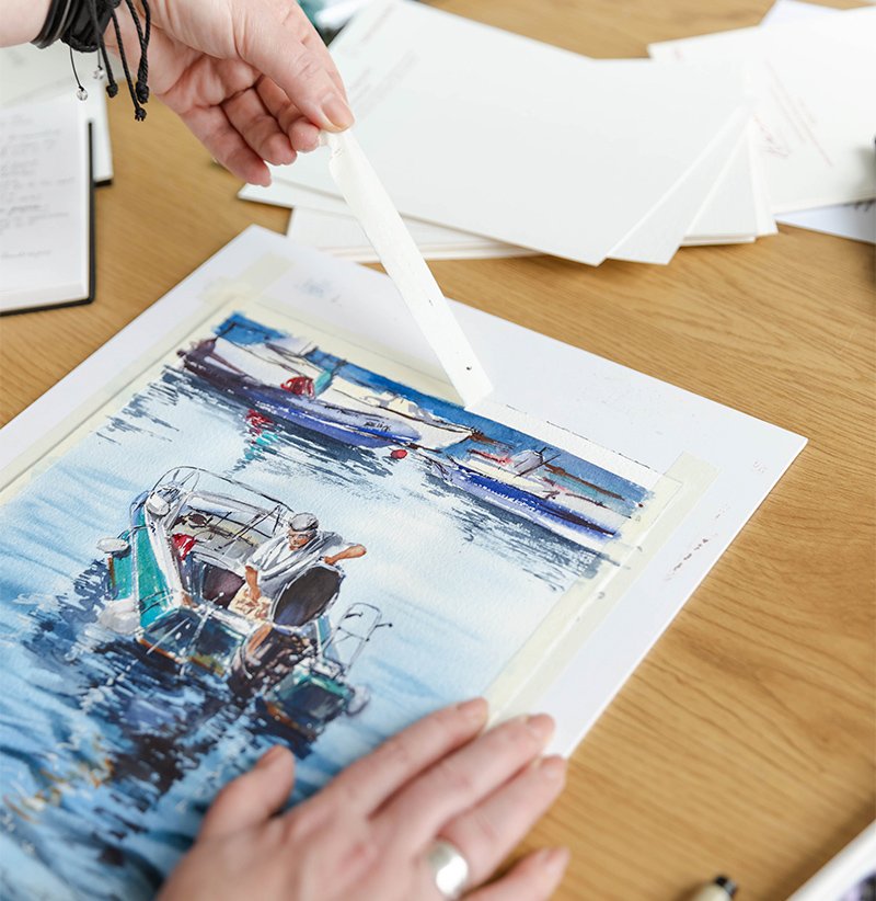Hands of watercolor artist Aliya Bodnar carefully taping the edges of a painting in her studio, preparing for a new creative session.