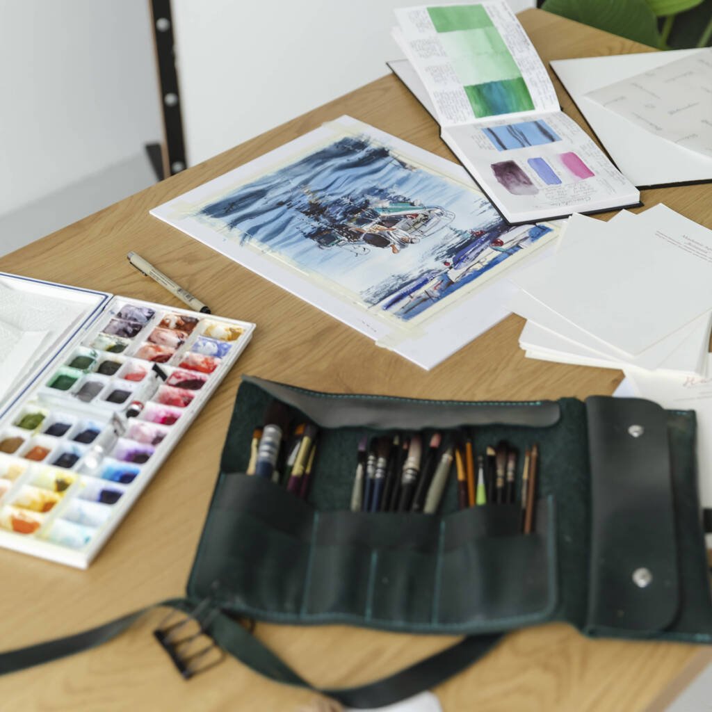 Flat lay of Aliya Bodnar’s watercolor workspace with brushes, palette, paper, and finished artworks.