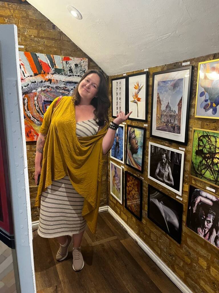 Artist Aliya Bodnar standing next to her watercolor paintings exhibited at The Vogue Seventh Edition Exhibition at Boomer Gallery, London.