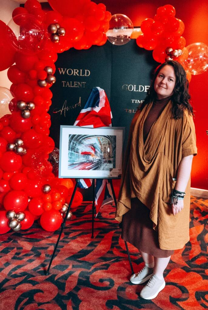 Aliya Bodnar standing next to her watercolor painting exhibited at the World Talent Award 2024 ceremony, wearing a brown coat against a red background.