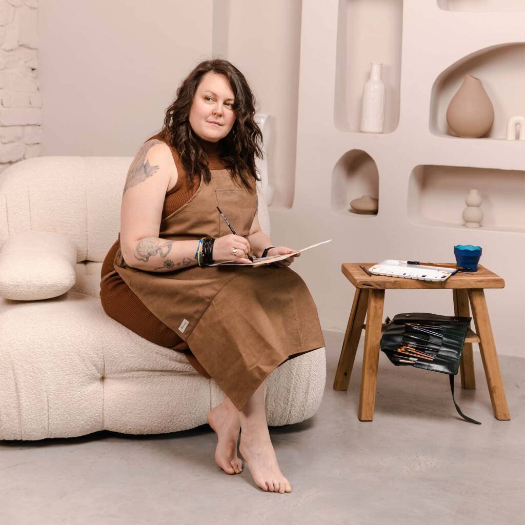 Aliya Bodnar seated in her art studio holding a framed watercolour painting, embracing her journey as a professional artist in the UK