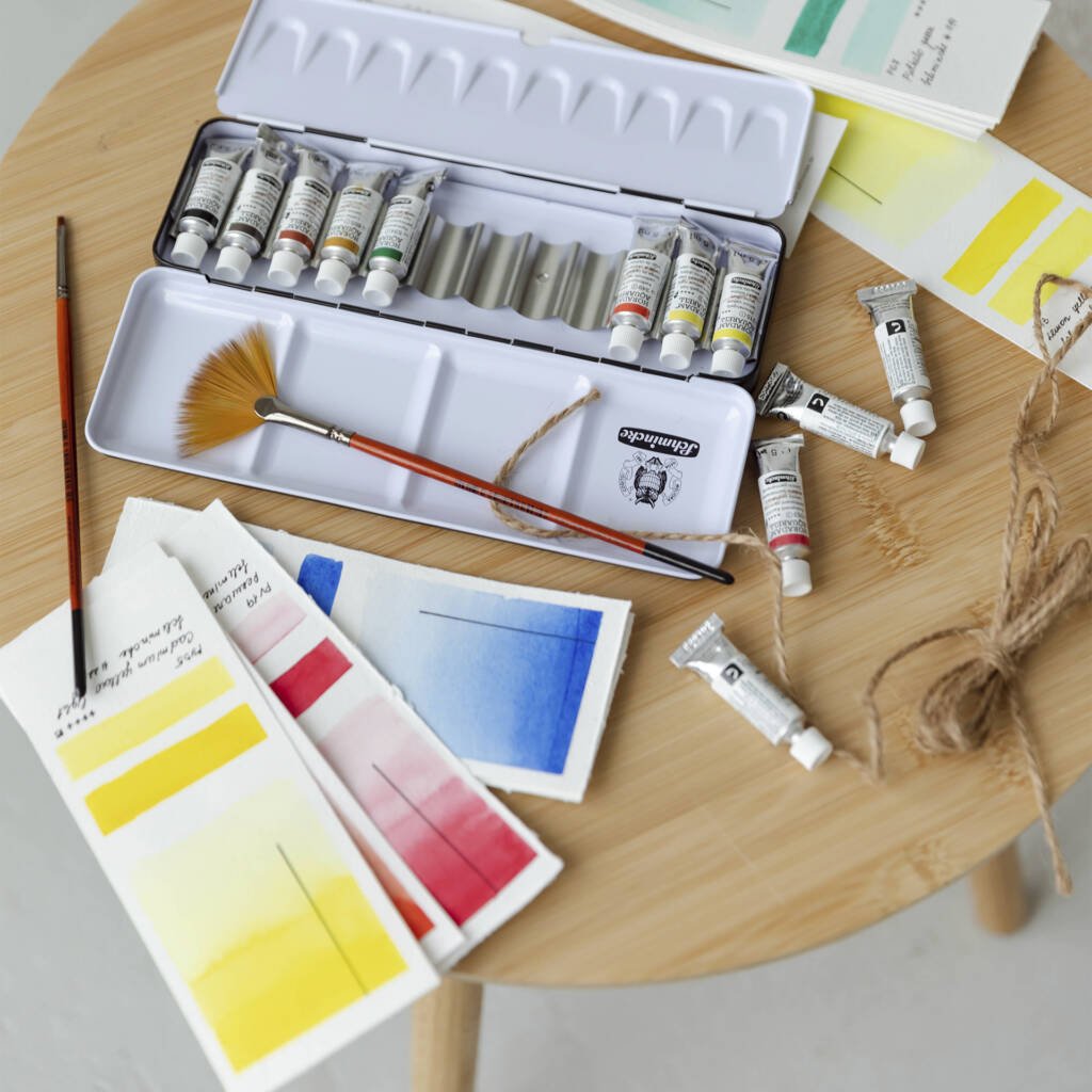 Flat lay of watercolor paintings, brushes, and art tools on a wooden table in Aliya Bodnar’s studio.