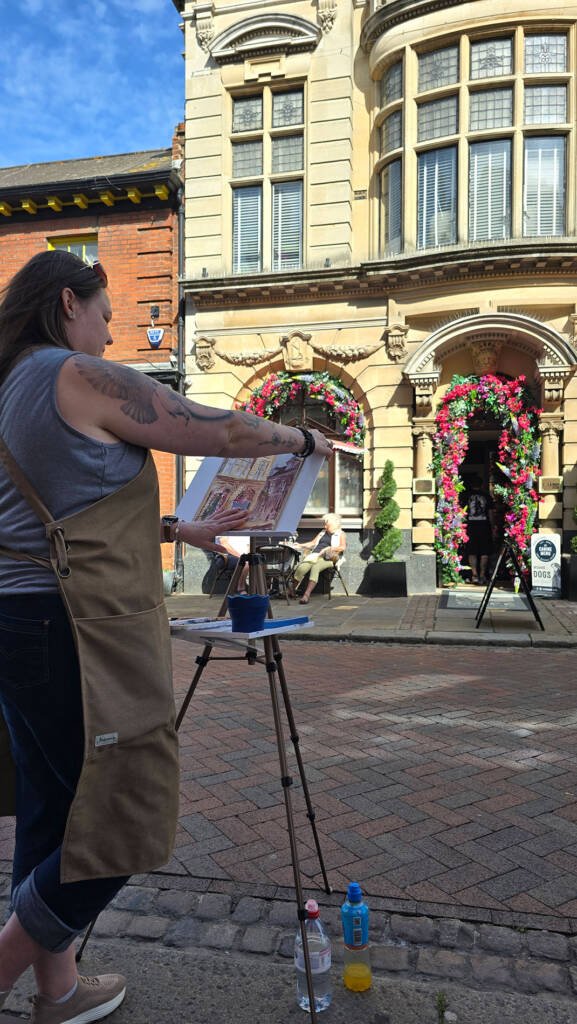 Aliya Bodnar painting en plein air during her solo art exhibition in Rochester, with historic architecture in the background.